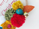 Colorful Christmas-themed decorative item with pom-poms and a bell on a white background.